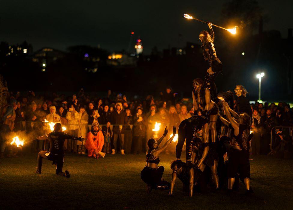 Acrobatics perform a stunt with fire in the foreground. In the background, rows of people watch on.