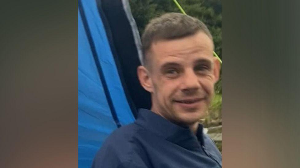 Rikki Berry posing for the camera, he is wearing a dark coloured polo neck shirt and appears to be in front of a tent and grass