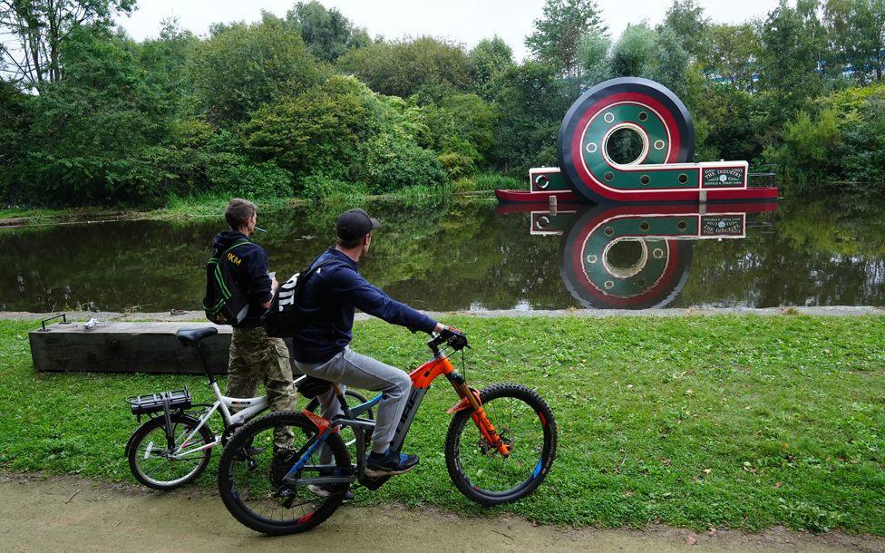 Two people on bicycles stop to look at the sculpture of a canal boat after installed on the Sheffield & Tinsley Canal, England.