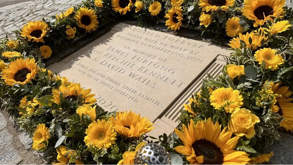 A picture of the stone memorial unveiled in 2023 shortly after its unfailing. Yellow flowers, including sunflowers, are included in a decoration around it. The plaque says: "United forever, never will we walk by without remembering you. In memory of: James Furlong, Joe Ritchie-Bennett and David Wails. James, Joe and David. Their families and all of those affected."