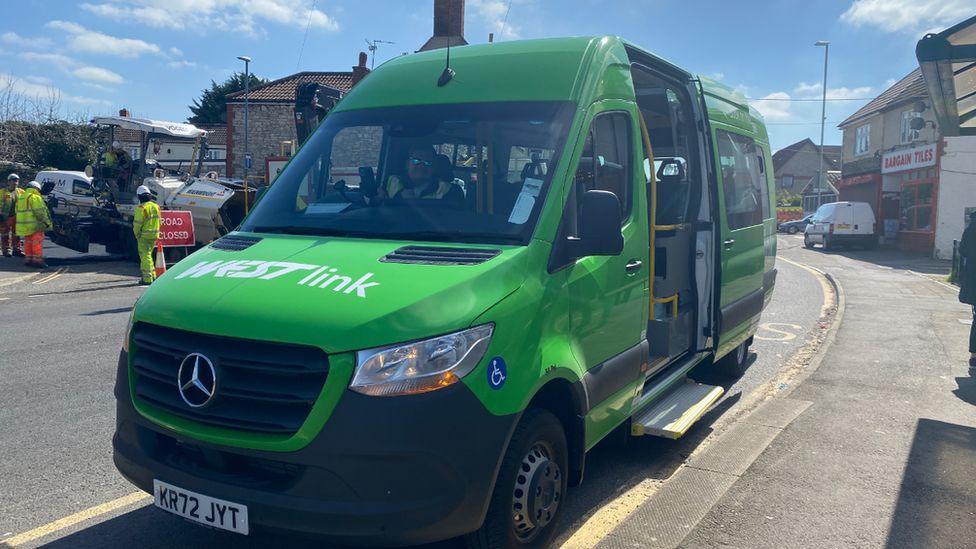 A green Westlink taxi van parked beside a pavement on a sunny day. It's side sliding door is open and there is a man wearing a hi-vis yellow vest sitting in the driver's seat. 