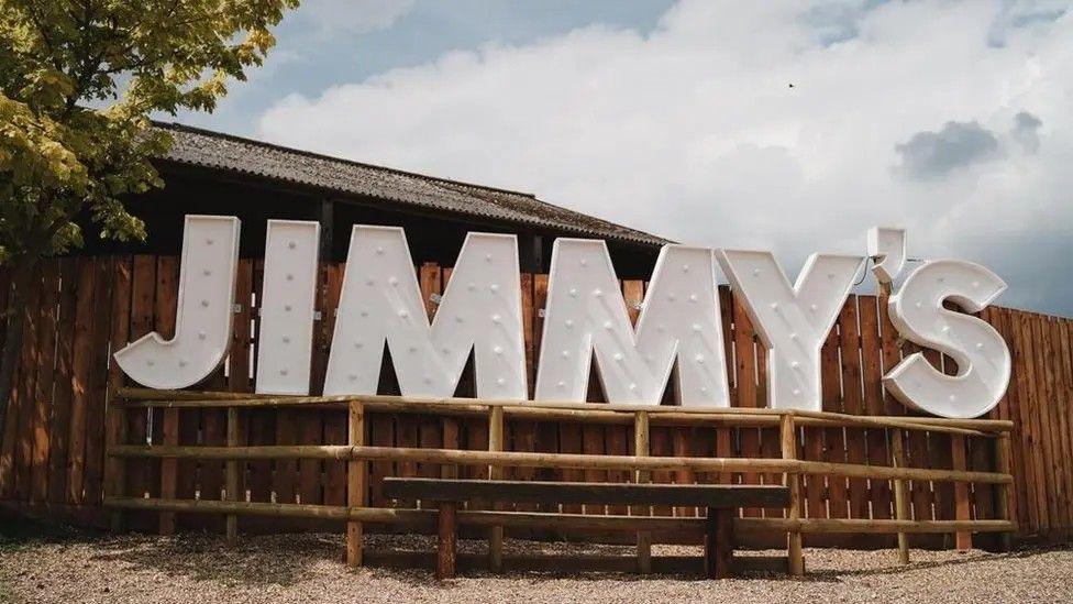 White letters with bulbs inside them spelling out Jimmy's. The letters are against a wooden fence on a raised platform at the farm. The picture has been taken during the day. 