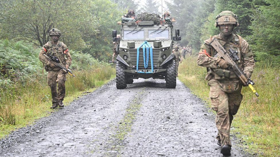 Two soldiers either side of a road, walking towards the camera. They are both armed with rifles and in camouflage with an armoured car between them in the middle distance.