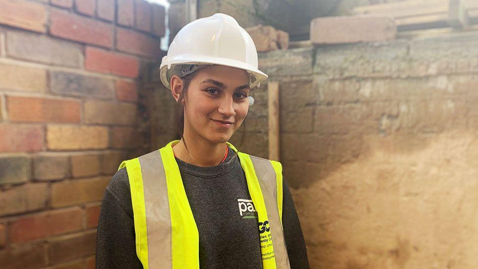 A young woman with dark hair in a white hard hat and yellow high visibility bib over a black jumper stands in front of a brick wall