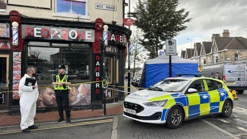 Police at the scene of the reported incident. The picture shows a police car in front of a blue police tent. There are two police officers in the picture - one wearing a police uniform and the other wearing a white hazmat suit. An area outside a shop called Red One Barbers is cordoned off with blue and white police tape.