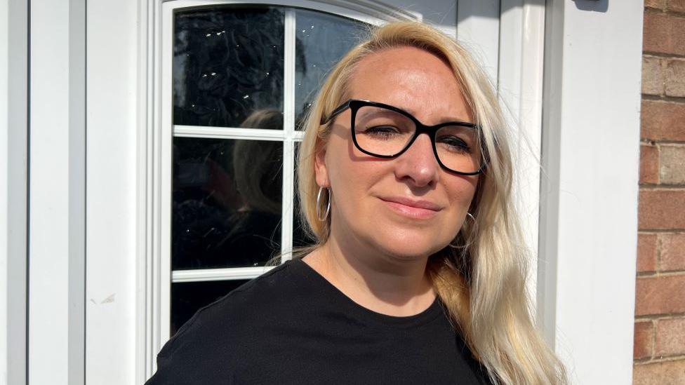 Dom Wolemder outside her home in Acomb, York. She wears a black top, black-rimmed glasses and is stood in front of her white door