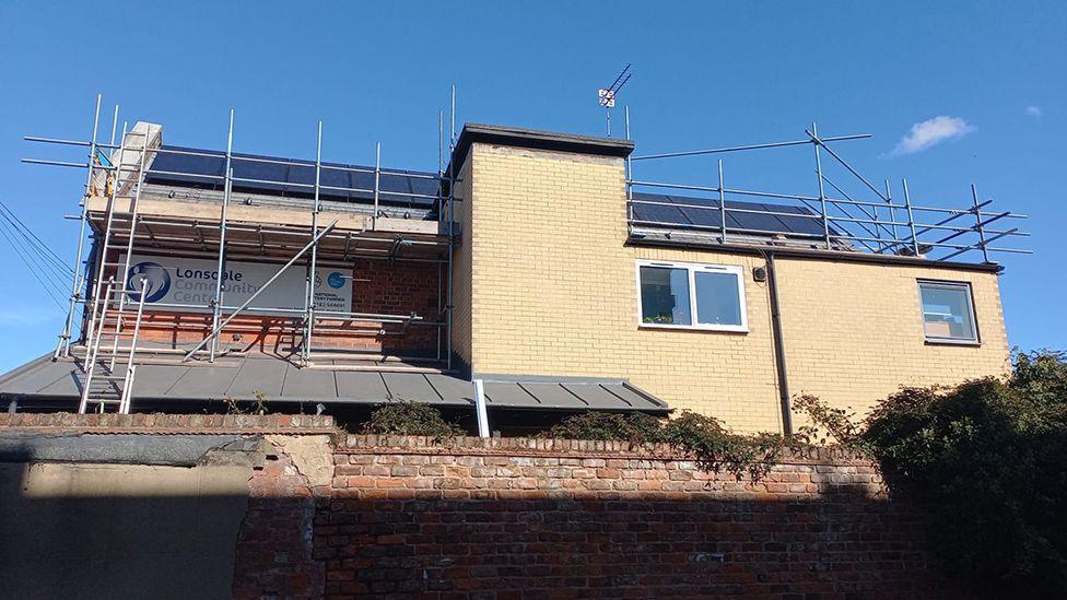 The solar panels being installed on the roof of the Lonsdale Community Centre a yellow brick building with a wall and hedges at the front. The building is surrounded by scaffolding.