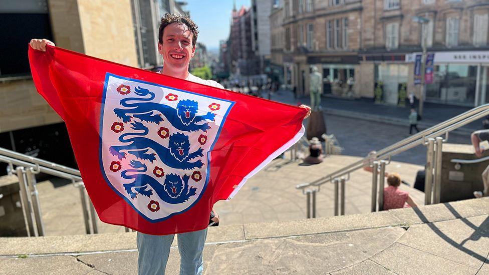 A man with dark hair poses with an red England football flag. He is standing at the top of a set of stone steps on a busy shopping street