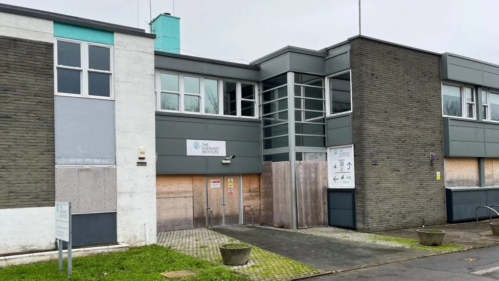 A two-storey school building in a 1960s-style, made of concrete, bricks and with faded coloured panels. The doors and some of the windows are boarded up.