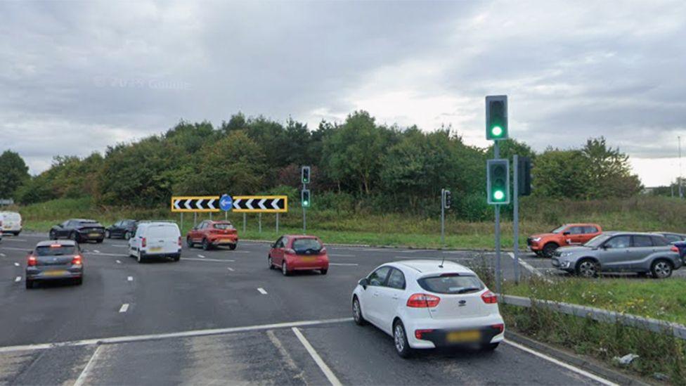 Moor Farm roundabout on the A19. The roundabout is covered by trees and grass. A queue of traffic is waiting to join the roundabout and other vehicles are crossing or waiting.