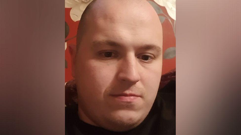 Darren Stevenson is a bald man wearing a dark top. He looks at the camera and appears to be in front of red patterned wallpaper