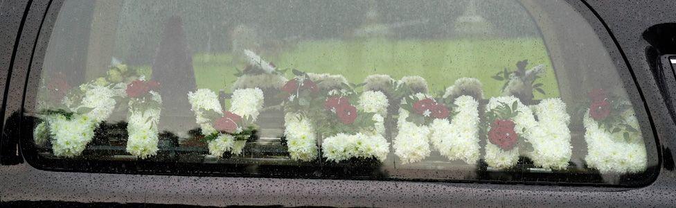 A floral tribute spelling "James" seen through a hearse window