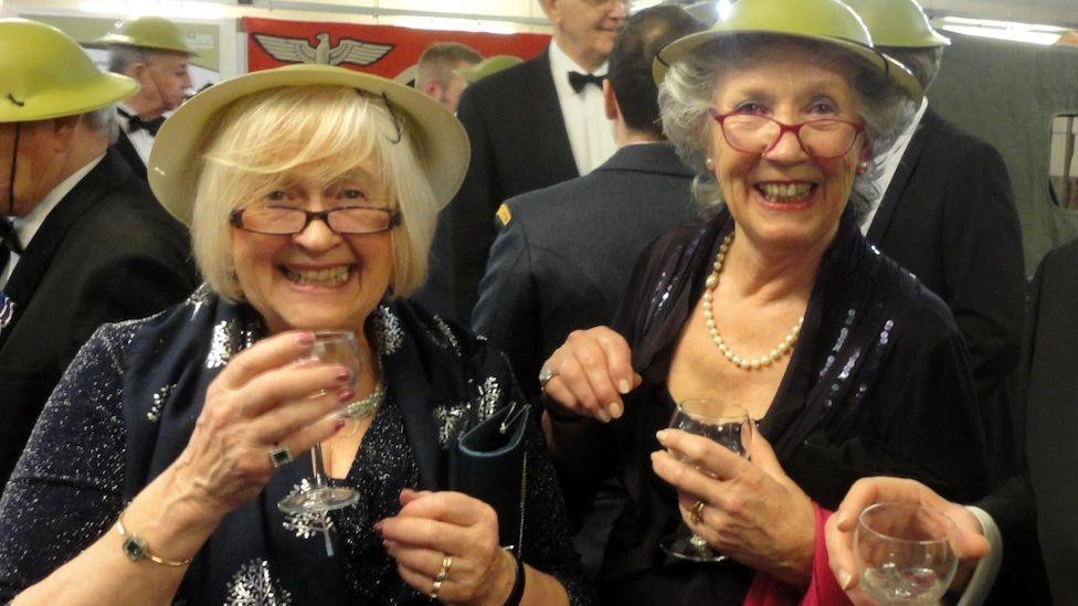 Penny Fell and her sister Juliet Francis. They are both wearing black evening dresses and yellow hats on their head, with wine glasses in their hands and smiling broadly. Behind them can be seen men in black tie. 