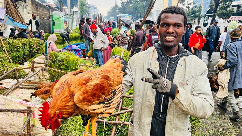 A young vendor at a market in Addis Ababa wearing gloves and holding up two fingers smiles at the camera as he grasps a live chicken in his other hand - Ethiopia, Wednesday 10 September 2025.