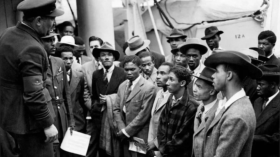 A black and white images shows a number of people gathered on board Empire Windrush, there is a dingy boat slightly covered in the background. The people are looking towards Johnny Smythe, who is holding a piece of paper and wearing his RAF uniform, standing next to another RAF officer who is talking to the group.