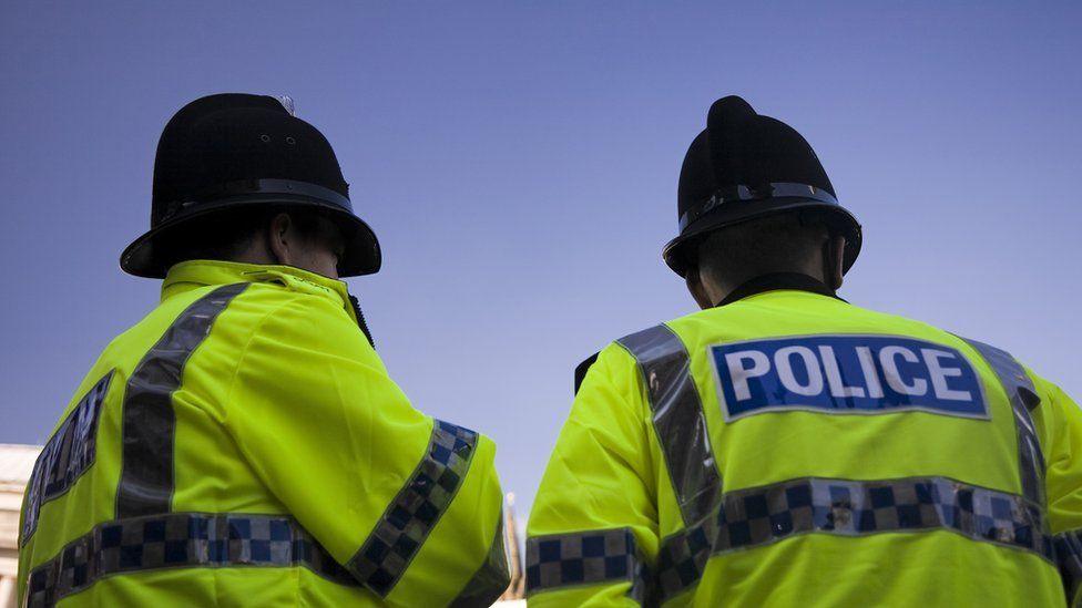 Stock image taken from the back of two police officers stood side by side wearing fluorescent police jackets and black police hats.