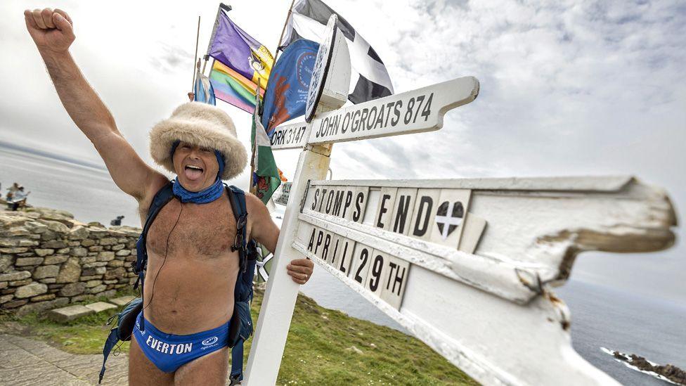 Speedo Mick wearing his blue Speedos, a woolly hat, scarf, backback and nothing else, with one arm around a white wooden direction sign at Land's End, and the other arm in the air and his mouth smiling and open. Pictured in 2023.