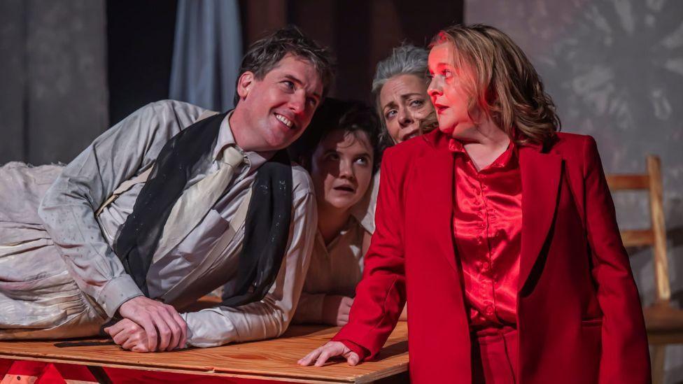 Still photography from Faust-ish play - a woman with blonde hair, in red silk pyjamas, leaning against a table. On the table a man with a grey suit is lying across it smiling. Beside her two women, both in grey bedwear, stand close to her, looking shocked. The staging is minimal, with several sheets draped across a red brick background.