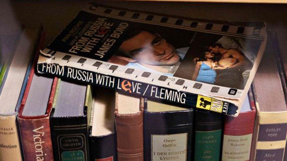 A battered paperback copy of From Russia with Love resting on a row of hardback books. The black spine has the title and Fleming written on it. Its cover shows Sean Connery from the film of the novel.