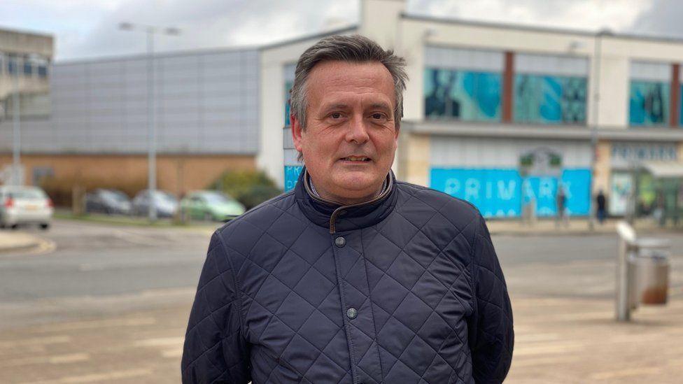 Dan Pickard with medium-length hair wearing a dark black padded jacket. He is standing on a pavement outside a two-storey Primark store with blue and white walls.