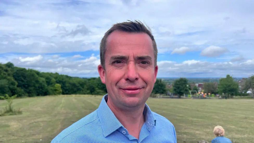 Calum Miller MP is showed posing for a photo outside with a large green field in the background. He is wearing a light blue smart shirt and has short brown hair.