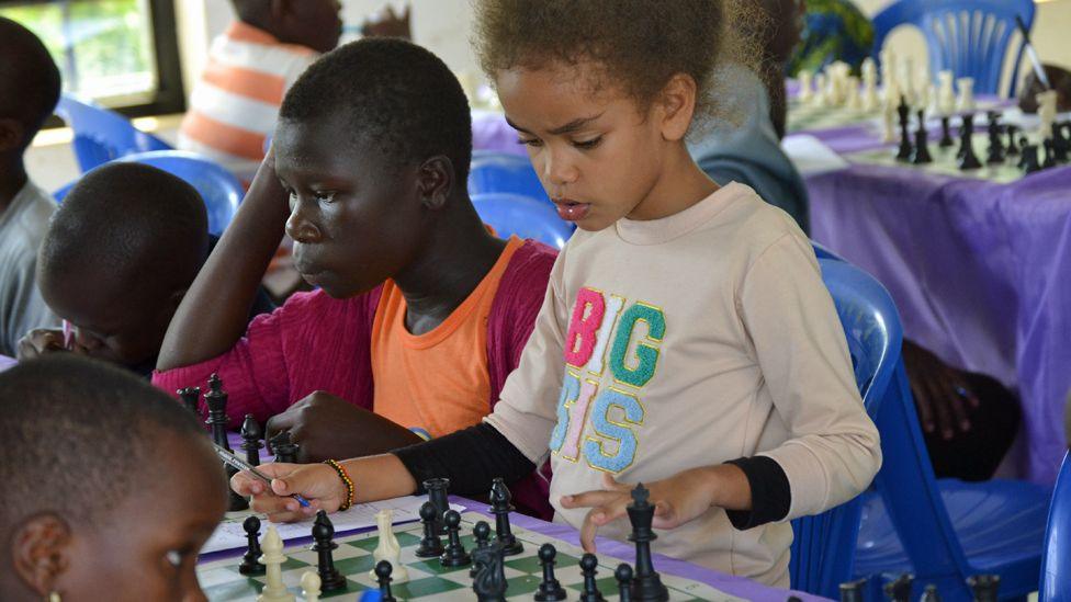 A young girl in a sweatshirt with the words 'Big Sis' on it stands at a chess board in a room where other chess games are being played by children. Next to her are some other players - all concentrating and looking down at the boards.