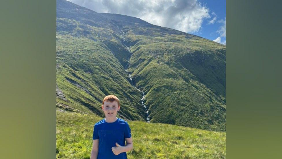 Alex standing in front of Ben Nevis. He had red hair and is wearing a blue T-shirt while giving a thumbs up to the camera. Behind him on the mountain, a stream runs along a valley down the grassy side of the mountain