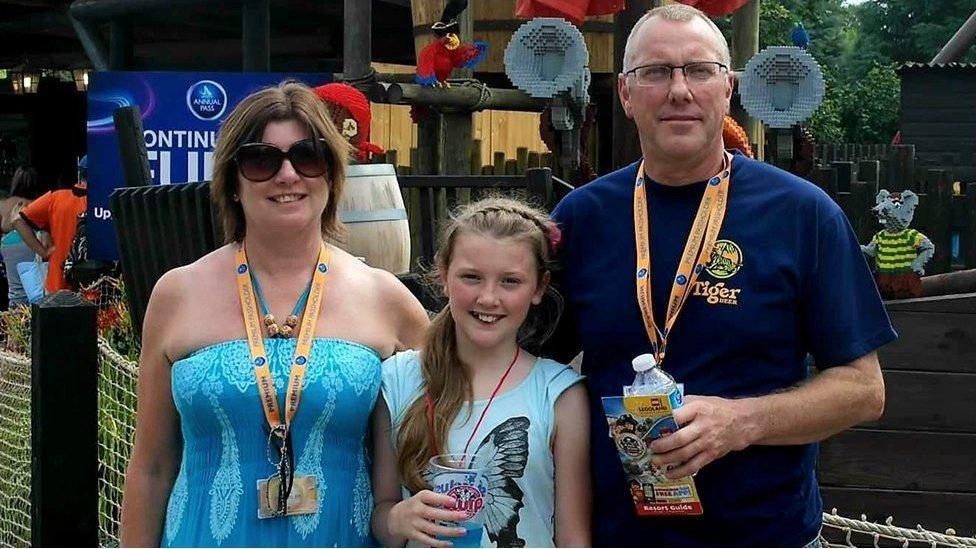 Jessica Lawson stands between her parents Brenda and Tony at a theme park. Jessica wears a blue top with butterfly design, Brenda wears a blue dress and dark sunglasses and Tony weaers a dark-blue T-shirt.