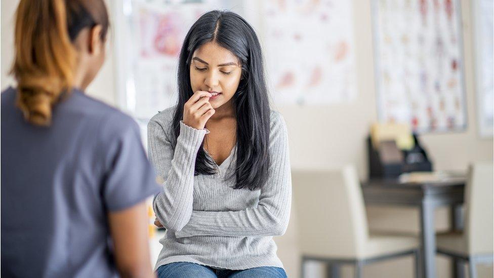 A Woman sat in a jumper and jeans in front of a health professional.