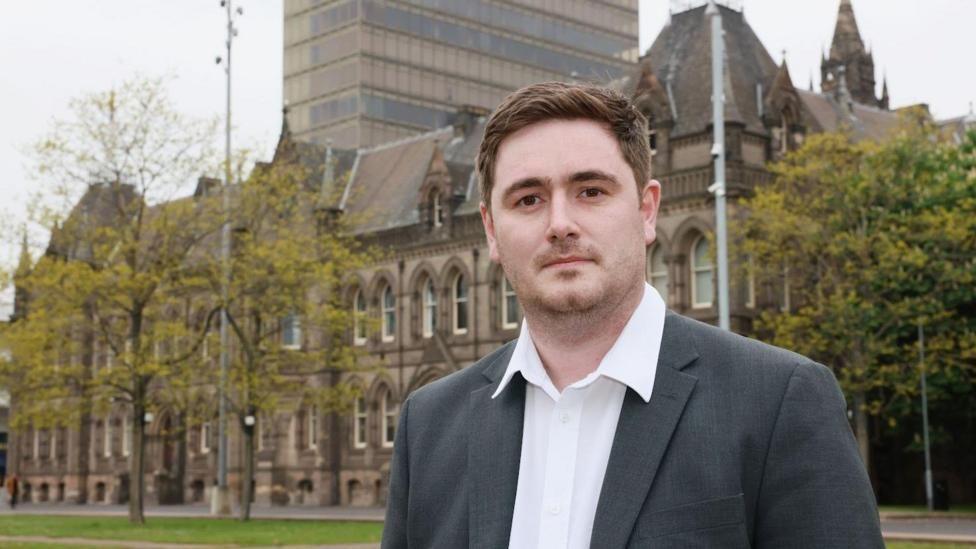 Chris Cooke looking into the camera. He's got brown, short hair and is wearing a grey blazer with white shirt. He's standing outside Middlesbrough Town Hall which is a large, gothic looking building.