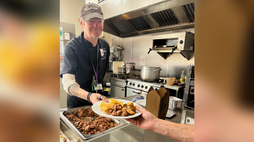 A man wearing chef uniform stands in a commercial kitchen. He smiles as he hands someone out of shot a plate of food.