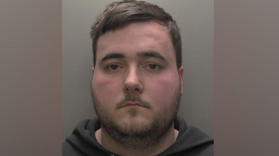 A police custody shot of a man with short brown hair and brown eyes. He has a thin stubbly beard and is wearing a black hooded sweatshirt.