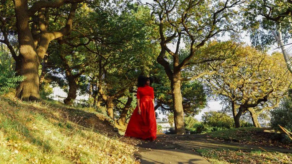 A black woman walking through a sunny park, filled with trees, in a bright red dress.