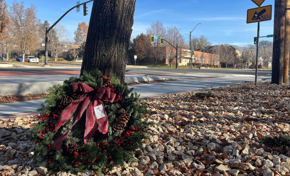 A christmas wreath with red berries and a tartan bow sits on the pebbled ground at the foot of a tree at a roadside, near a yellow traffic sign on a bright blue-skyed day.