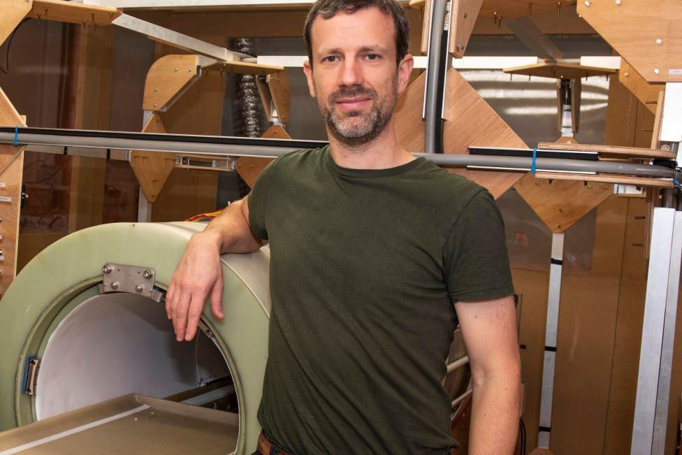 Man - Dr Lionel Broche - in a green T-shirt, smiling and looking at camera. his arm is resting on a green scanner machine.