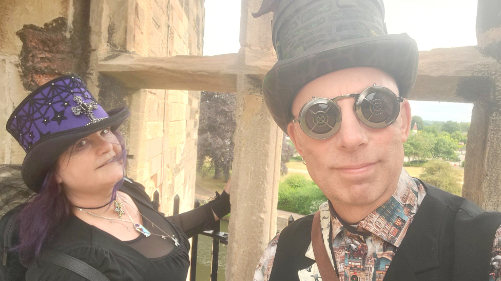 June Gothika is on the left she is wearing a black dress with some silver jewellery at her neck and a blue and black top hat while Gary Gimmick is wearing round steampunk black glasses and a black vest over a patterned shirt. They are standing against a stone window at Newark Castle.