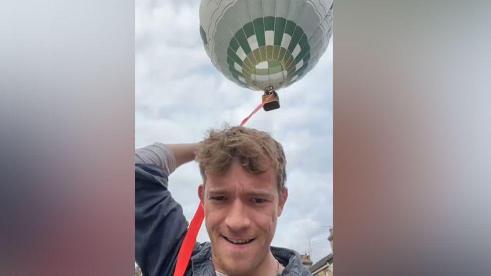 Eben Chinh, holding into a red balloon guide rope, over his shoulder, looking at the camera. He is wearing a blue top and has short fair hair. A hot air balloon is above him.