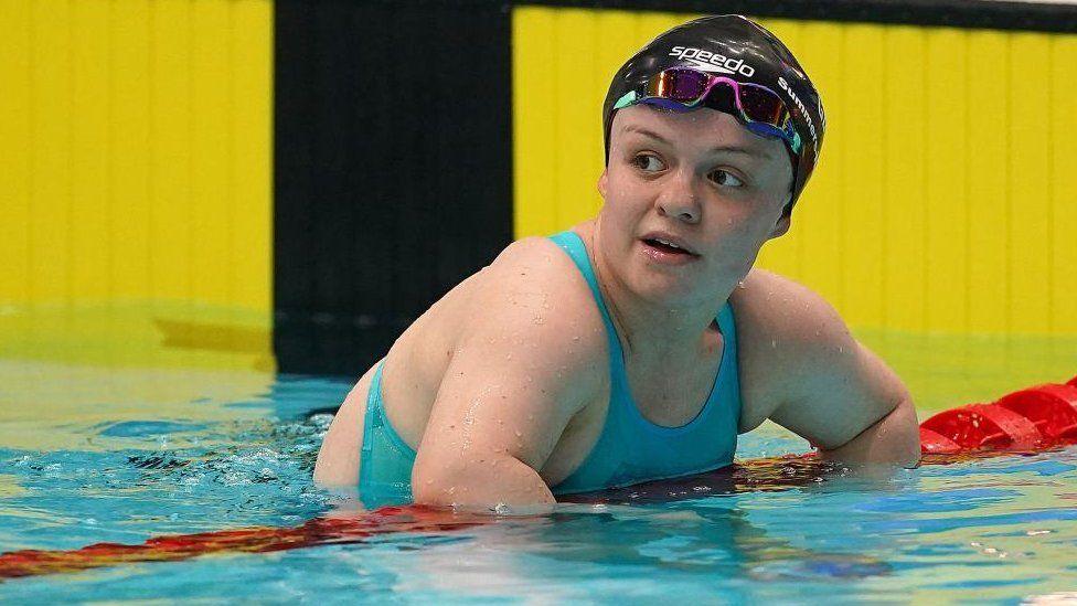 Maisie Summers-Newton with a black swimming cap and blue goggles, wearing a blue swimming costume and leaning over a red lane rope in a yellow-surround swimming pool