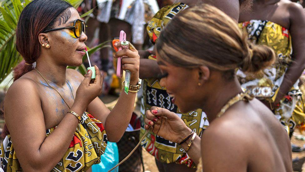 A side profile of a Calabar carnival participant uses a mobile phone as a mirror to apply her lipstick