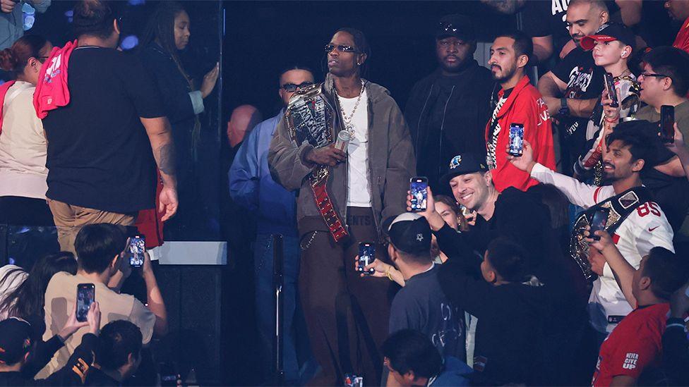 Travis Scott enters the arena during Monday Night RAW, holding a belt, wearing dark grey tracksuits and a white top, coming through fans.