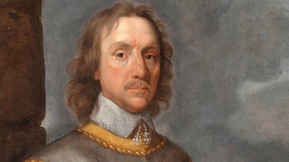 An artist's painting of Oliver Cromwell. He has long brown hair and is wearing a collared grey tunic.