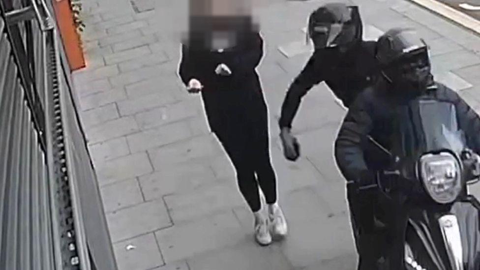 Two people riding a moped dressed in black and wearing helmets snatch a phone from a woman, whose face is blurred in the image, on the pavement