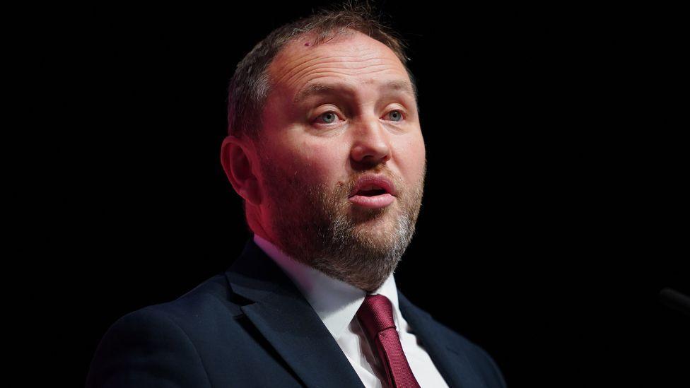 Ian Murray, with dark receding hair and a grey and ginger beard, photographed in a medium close-up shot. He is in front of a dark background and wearing a dark suit, white shirt and red tie