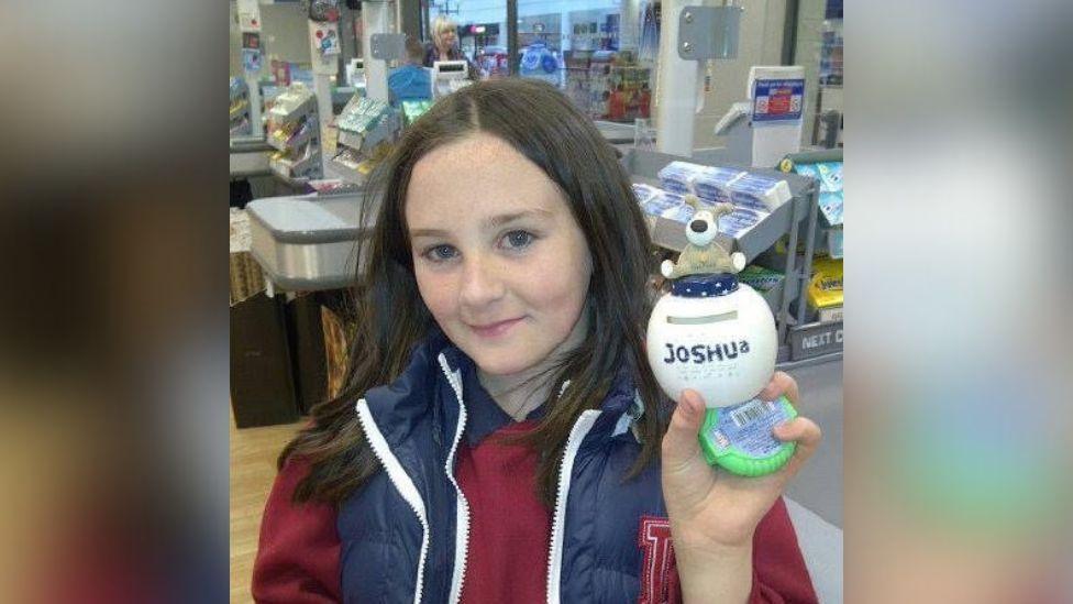 Joshua as a young boy. He wears his hair long and is smiling at the camera. He is wearing a red sweater and a blue body warmer.  He is holding a small white ball which has his name written on it. The photo is taken beside the checkout in a small shop. 