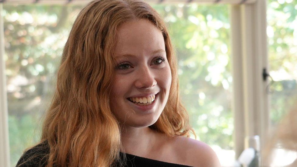 A close-up of Roxy Longworth, who had long red hair and is wearing a black off-the shoulder jumper and is looking to the right and smiling. Behind her is a window. 