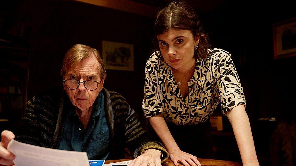 Timothy Spall, as character John Chapel, seated at a table holding a document with Gwyneth Keyworth, playing Det Sg Janie Mallowen, standing leaning over next to him. Both look at the camera with interrogative expressions.