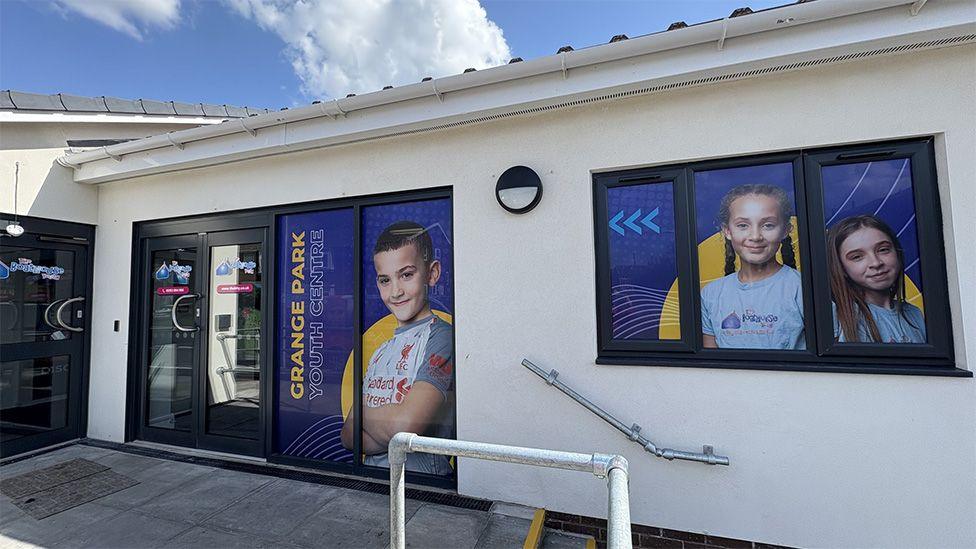 The entrance to Grange Park Youth Centre, a single storey, white rendered building with large black-framed windows and blue doors that have full length photos of smiling young people.