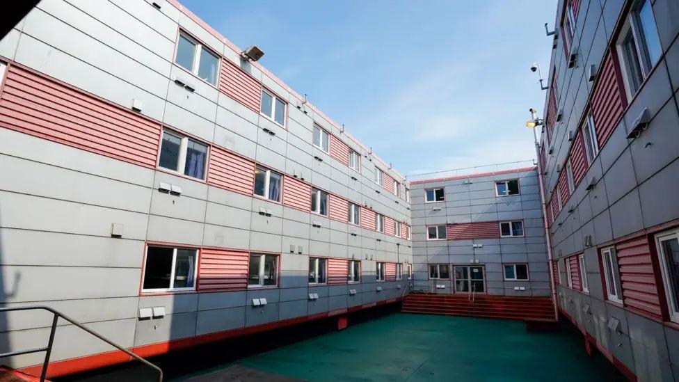 The interior courtyard on board the Bibby Stockholm. Grey and red walls of three stories in height surround a narrow, green, deck.