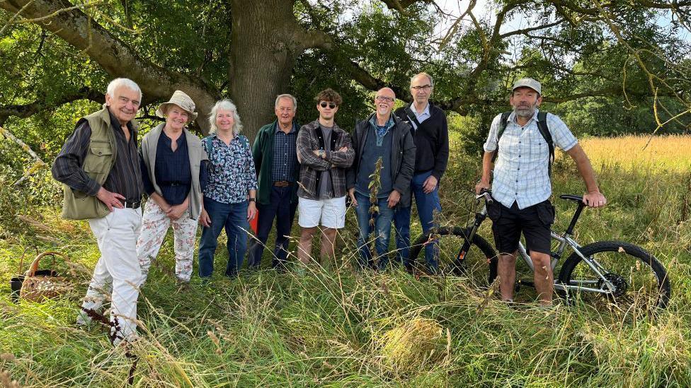 Eight people stand in a field under a tree