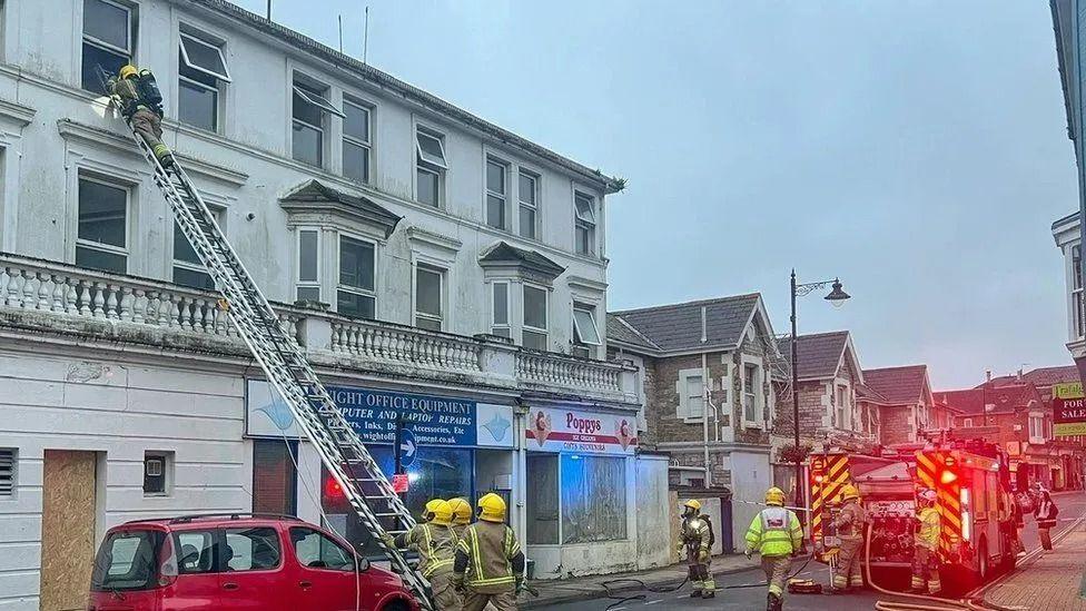 A ladder rests against a third floor window of the hotel with a firefighter looking into the window firefighters and a fire engine in the road below